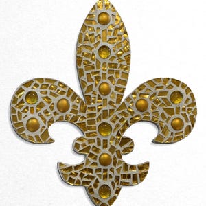 May include: A gold fleur-de-lis wall decor made of mosaic tiles with a white background. The fleur-de-lis is decorated with small, round, gold and yellow glass beads.