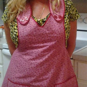 May include: A pink floral print apron with red trim and two large pockets. The apron has a V-neck and a collar with two buttons.