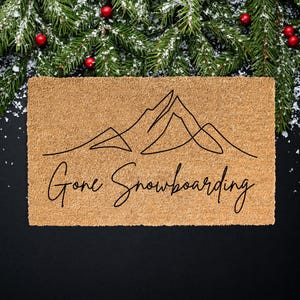 May include: A rectangular doormat made of natural coir fiber. The mat features a minimalist black line drawing of mountain peaks and the cursive text "Gone Snowboarding". Festive greenery and red berries decorate the top.