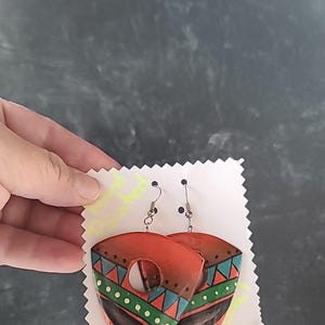 May include: A pair of orange, green, and blue geometric patterned earrings with silver hooks. The earrings are shaped like a teardrop with a hole in the center.