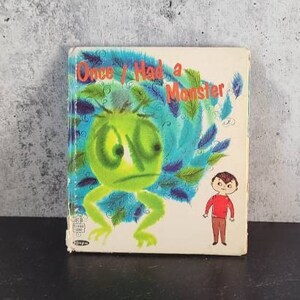 Puede incluir: Una portada de libro infantil vintage con un monstruo verde con plumas azules y un niño con una camisa roja. El título del libro es "Once I Had a Monster."