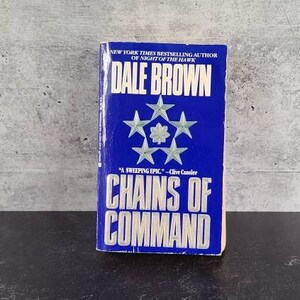 May include: A paperback book titled "CHAINS OF COMMAND" by Dale Brown. The cover is blue with white text and star graphics. The book is a "New York Times Bestselling Author" novel.