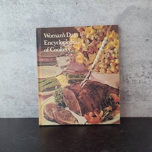May include: A vintage cookbook titled "Woman's Day Encyclopedia of Cookery" with a photo of a roasted meat dish on the cover.