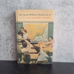 May include: A vintage book titled "The Rand McNally Handbook of MAP AND GLOBE USAGE". The cover features a classroom scene with students and a teacher looking at a map of the United States.