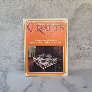 May include: A book titled "The Complete Encyclopedia of Crafts" with an orange and yellow cover. The book features a photo of a colourful patchwork cushion on a wooden chair.