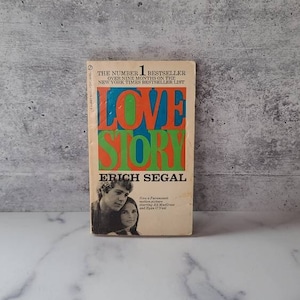 以下が含まれることがあります： エリック・シーガルによる小説「ラブストーリー」のペーパーバック版。表紙には、赤、オレンジ、緑のタイトルと、若いカップルの写真が載っています。