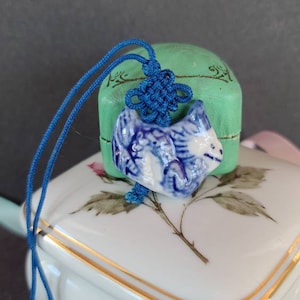 May include: A blue and white ceramic pendant of a stylized animal, possibly a cat or dog, with a blue cord tied in a knot. The pendant is sitting on a white porcelain box with a gold trim.