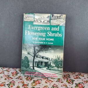 May include: A vintage book titled "Evergreen and Flowering Shrubs for Your Home" by Katharine M-P. Cloud. The book has a green cover with images of houses and trees. It rests on a floral patterned surface.