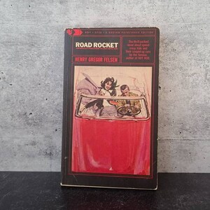 May include: A vintage paperback book cover with a red, white, and black color scheme. The cover features an illustration of a red car with a couple inside. The title of the book is "Road Rocket" by Henry Gregor Felsen.