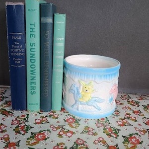 Puede incluir: Un macetero de cerámica azul con un diseño de carrusel que presenta un conejito amarillo, un caballo rosa y un caballo azul. El macetero está sentado en una mesa con un patrón floral.