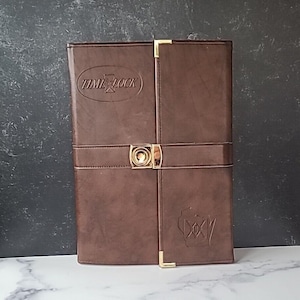 Puede incluir: Un diario encuadernado en cuero marrón con detalles dorados y las palabras "Time Lock" en relieve en la portada. El diario también tiene un cierre dorado y un diseño dorado en relieve en la contraportada.
