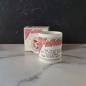 May include: A white ceramic coffee mug with a green, pink, and blue design. The mug features a cartoon illustration of a woman and the text "The fear that you'll raise your children the right way..."