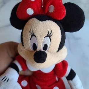 K&ouml;nnte beinhalten: Pl&uuml;schpuppe Minnie Mouse mit rotem Kleid mit wei&szlig;en Punkten, passender Schleife mit wei&szlig;en Punkten und schwarzen Ohren. Die Puppe hat ein cremefarbenes Gesicht mit gestickten Augen und einer schwarzen Nase. Die Arme sind wei&szlig; mit schwarzen Streifen.