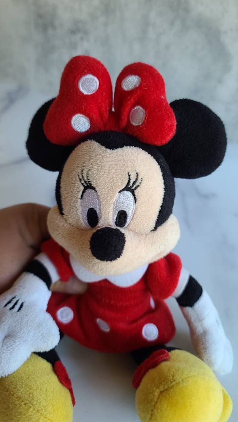 K&ouml;nnte beinhalten: Pl&uuml;schpuppe Minnie Mouse mit rotem Kleid mit wei&szlig;en Punkten, passender Schleife mit wei&szlig;en Punkten und schwarzen Ohren. Die Puppe hat ein cremefarbenes Gesicht mit gestickten Augen und einer schwarzen Nase. Die Arme sind wei&szlig; mit schwarzen Streifen.