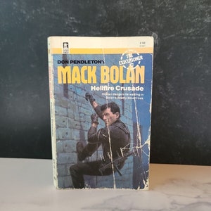 May include: A paperback book cover featuring a man hanging from a wall with a gun in his hand. The book title is "Mack Bolan: Hellfire Crusade" and the author is Don Pendleton. The book is part of "The Executioner" series.