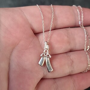 May include: A silver-colored necklace featuring a pendant with a Celtic knot design and two ribbon-like elements. The necklace is displayed against a neutral background, highlighting the intricate details of the pendant and chain.