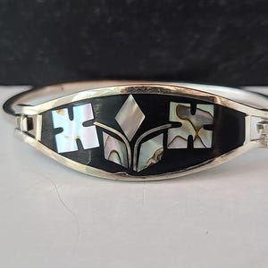 May include: Silver-tone bracelet with a black center panel. The panel features inlaid mother-of-pearl and abalone shell designs. The bracelet has a hinged clasp for easy wear.