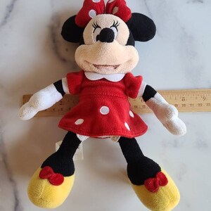 K&ouml;nnte beinhalten: Ein Pl&uuml;schtier von Minnie Mouse, mit einem roten Kleid mit wei&szlig;en Punkten und einer passenden Schleife. Die Puppe hat schwarze Ohren, ein beiges Gesicht und gelbe Schuhe mit roten Schleifen. Die Puppe ist etwa 25 cm gro&szlig;.