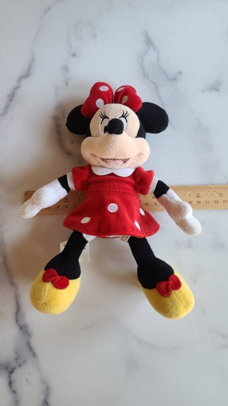 K&ouml;nnte beinhalten: Ein Pl&uuml;schtier von Minnie Mouse, mit einem roten Kleid mit wei&szlig;en Punkten und einer passenden Schleife. Die Puppe hat schwarze Ohren, ein beiges Gesicht und gelbe Schuhe mit roten Schleifen. Die Puppe ist etwa 25 cm gro&szlig;.