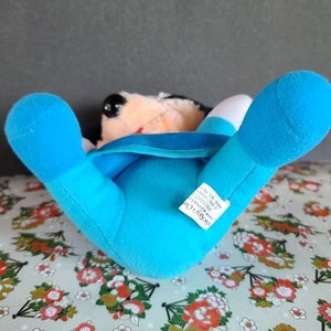 May include: Blue plush Mickey Mouse toy lying on its back with its arms and legs outstretched. The toy has a white face with black eyes and a red mouth. The toy is wearing a blue shirt with a white collar. The toy has a tag that reads "Mickey's Club", "The Mouse with the Magic", "All New", "Disney", "Made in China".