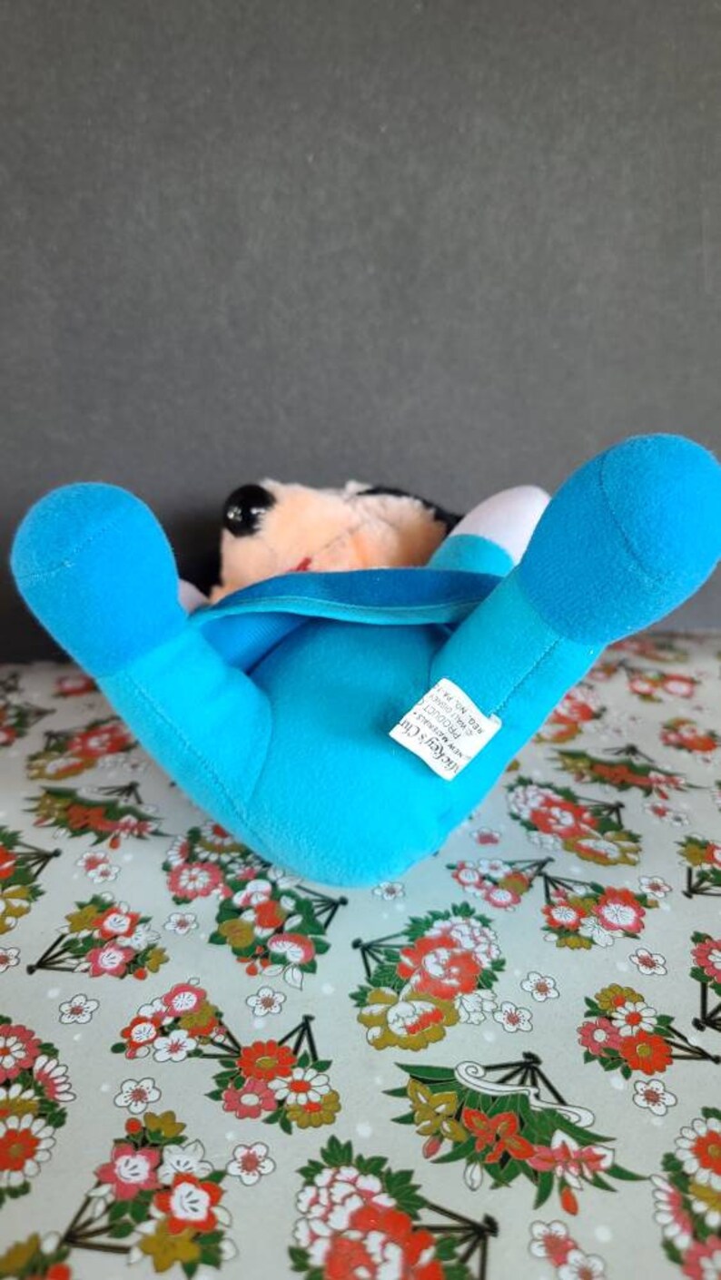 May include: Blue plush Mickey Mouse toy lying on its back with its arms and legs outstretched. The toy has a white face with black eyes and a red mouth. The toy is wearing a blue shirt with a white collar. The toy has a tag that reads "Mickey's Club", "The Mouse with the Magic", "All New", "Disney", "Made in China".