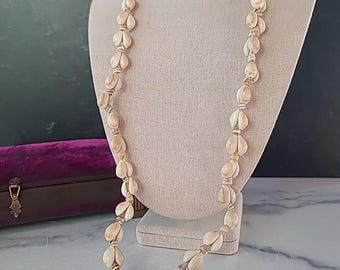 Puede incluir: Un collar largo hecho de conchas de color crema, exhibido en un maniquí. Las conchas están ensartadas con un cordón dorado. El collar es una pieza vintage. El fondo incluye una caja de terciopelo púrpura.