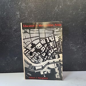 以下が含まれることがあります： ロバート・J・ハヴィガースト著「Education in Metropolitan Areas」というタイトルの本。表紙には、都市の白黒地図が描かれています。本は暗い背景に対して直立しています。