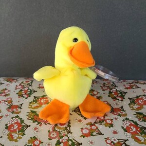 May include: A plush yellow duck with an orange beak and feet. The duck has black eyes and small wings. It is sitting on a floral patterned surface. A clear tag is attached to the duck.