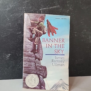 May include: A vintage paperback book titled "Banner in the Sky" by James Ramsey Ullman. The cover shows a climber ascending a rock face with a red flag. The book has a Scholastic logo and a Newbery Honor seal.