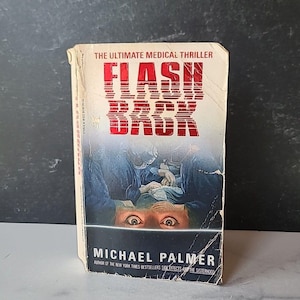 May include: A paperback book titled "Flash Back" by Michael Palmer, described as "The Ultimate Medical Thriller". The cover features a close-up of a pair of wide-open eyes, with a surgical scene in the background.