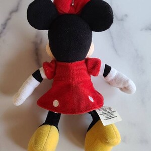 K&ouml;nnte beinhalten: Eine Pl&uuml;schpuppe von Minnie Mouse, mit schwarzem Kopf, gro&szlig;en Ohren und einer roten Schleife. Sie tr&auml;gt ein rotes Kleid mit wei&szlig;en Punkten, wei&szlig;e Handschuhe und gelbe Schuhe. Die Puppe ist etwa 20 cm gro&szlig;.