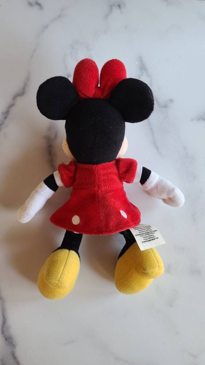 K&ouml;nnte beinhalten: Eine Pl&uuml;schpuppe von Minnie Mouse, mit schwarzem Kopf, gro&szlig;en Ohren und einer roten Schleife. Sie tr&auml;gt ein rotes Kleid mit wei&szlig;en Punkten, wei&szlig;e Handschuhe und gelbe Schuhe. Die Puppe ist etwa 20 cm gro&szlig;.