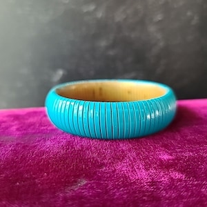 May include: A turquoise blue bangle bracelet with vertical grooves. The inside of the bracelet is a light wood color. The bracelet is resting on a pink velvet surface.