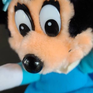 May include: A plush toy of a cartoon mouse with black fur, large eyes, and a blue bow. The mouse is wearing a blue dress and has a white collar.