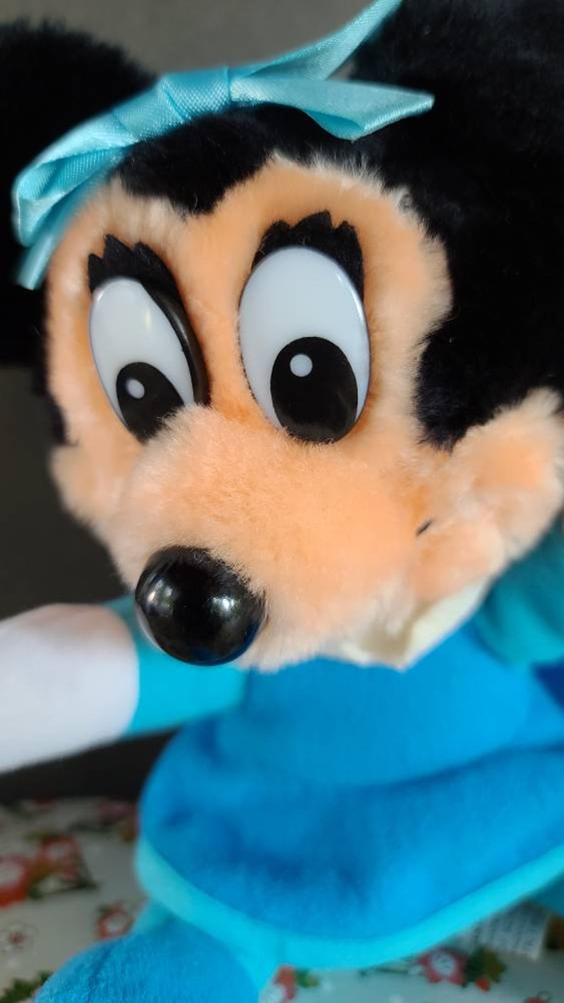 May include: A plush toy of a cartoon mouse with black fur, large eyes, and a blue bow. The mouse is wearing a blue dress and has a white collar.