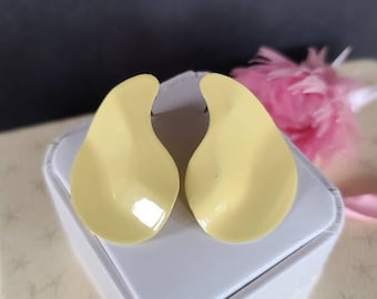 May include: A pair of pale yellow, teardrop-shaped earrings. The earrings are smooth and glossy, with a slight curve. They are displayed on a white surface, with a soft pink flower in the background. The earrings are a simple, elegant design.