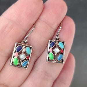 May include: A pair of silver-toned rectangular earrings. Each earring features a mosaic of colorful gemstones in shades of blue, green, pink, white, and red. The earrings have a simple hook closure.