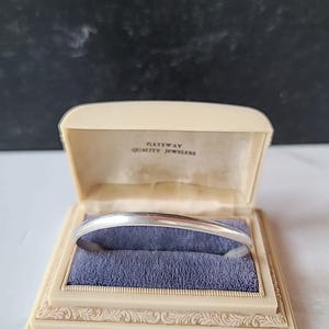 May include: A silver bangle bracelet displayed in an open, vintage jewelry box. The box is cream-colored with a blue velvet interior and the text "GATEWAY QUALITY JEWELERS" printed on the inside of the lid.
