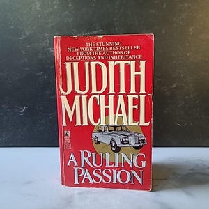 以下が含まれることがあります： ジュディス・マイケルの「A Ruling Passion」というタイトルのヴィンテージペーパーバック。表紙は赤色で白い文字で、クラシックカーのイラストが描かれています。この本はニューヨーク・タイムズのベストセラーです。