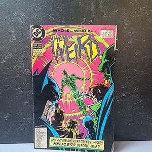 Puede incluir: Un cómic vintage titulado "The Weird" con una portada vibrante que presenta una figura verde, Batman y otros personajes. La portada tiene un fondo rosa y amarillo con el texto "Who is... What is..." y "And why are America's greatest heroes helpless before him?"