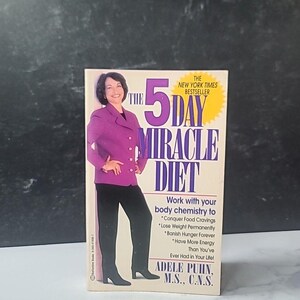 Puede incluir: Un libro de bolsillo titulado "The 5 Day Miracle Diet" de Adele Puhn. La portada presenta a una mujer con una blazer morada y pantalones negros. El libro promete ayudar con los antojos de comida, la pérdida de peso y la energía.