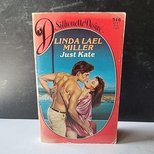 Puede incluir: Libro de bolsillo vintage titulado "Just Kate" de Linda Lael Miller, parte de la serie Silhouette Desire. La portada presenta una ilustración romántica de una pareja abrazándose. El libro tiene un borde rojo y el logotipo de la serie.