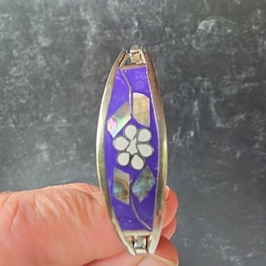 Peut inclure: Bracelet argenté avec incrustation violette ornée d'une fleur blanche et d'incrustations nacrées. Le bracelet de forme ovale est doté d'un fermoir à charnière. Le design est symétrique et les couleurs sont vives.