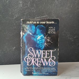 May include: A paperback book cover with a photo of a couple dancing. The title of the book is "Sweet Dreams" and the text on the cover says "The love and life story of Patsy Cline by the author of Coal Miner's Daughter, George Vecsey, with Leonore Fleischer. Now a major motion picture."