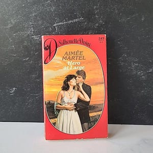 May include: A red paperback book with the title "Hero at Large" by Aimee Martel. The book is part of the Silhouette Desire series, number 249.