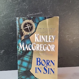 May include: A paperback book titled "Kinley Macgregor Born in Sin" with a gold and blue cover. The cover features a tartan pattern and a decorative emblem. The author's name is Stephanie Laurens.