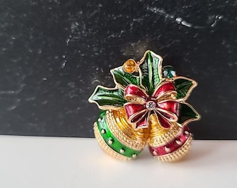 May include: A gold-toned Christmas brooch featuring two bell-shaped ornaments with red and green enamel accents. A red bow with a crystal center and green holly leaves complete the festive design. The brooch is a holiday-themed accessory.