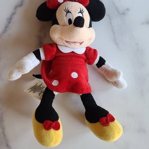 K&ouml;nnte beinhalten: Eine Pl&uuml;schpuppe von Minnie Mouse. Die Puppe hat ein rotes Kleid mit wei&szlig;en Punkten, eine passende Schleife und gelbe Schuhe. Die Puppe hat schwarze Ohren und ein beiges Gesicht. Die Arme und H&auml;nde der Puppe sind wei&szlig;.