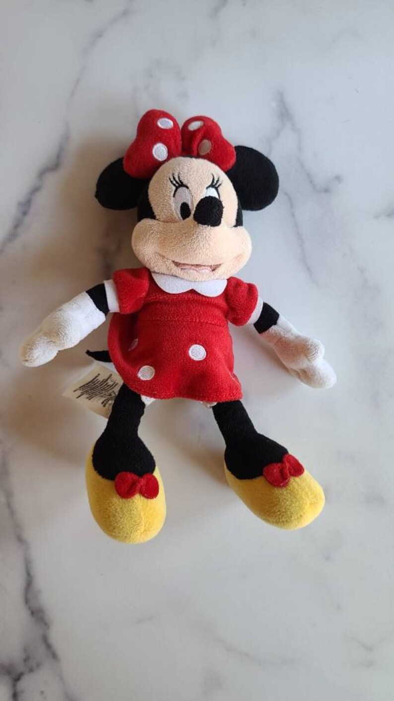 K&ouml;nnte beinhalten: Eine Pl&uuml;schpuppe von Minnie Mouse. Die Puppe hat ein rotes Kleid mit wei&szlig;en Punkten, eine passende Schleife und gelbe Schuhe. Die Puppe hat schwarze Ohren und ein beiges Gesicht. Die Arme und H&auml;nde der Puppe sind wei&szlig;.