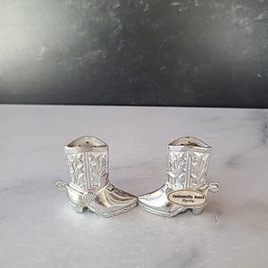 May include: Two silver-colored cowboy boot salt and pepper shakers. Each boot has decorative floral patterns and a small spur on the back. One shaker has a label that reads "Jacksonville Beach Florida."